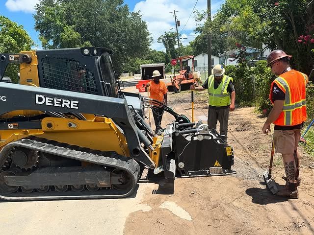 Road construction: John Deere skid steer milling asphalt. Three workers observe; one in orange vest.