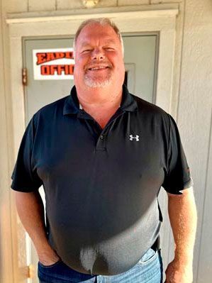 Man in black polo shirt and jeans smiles outside a door.