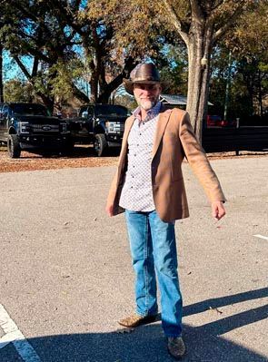Man in tan jacket and hat stands outdoors in front of two black trucks.
