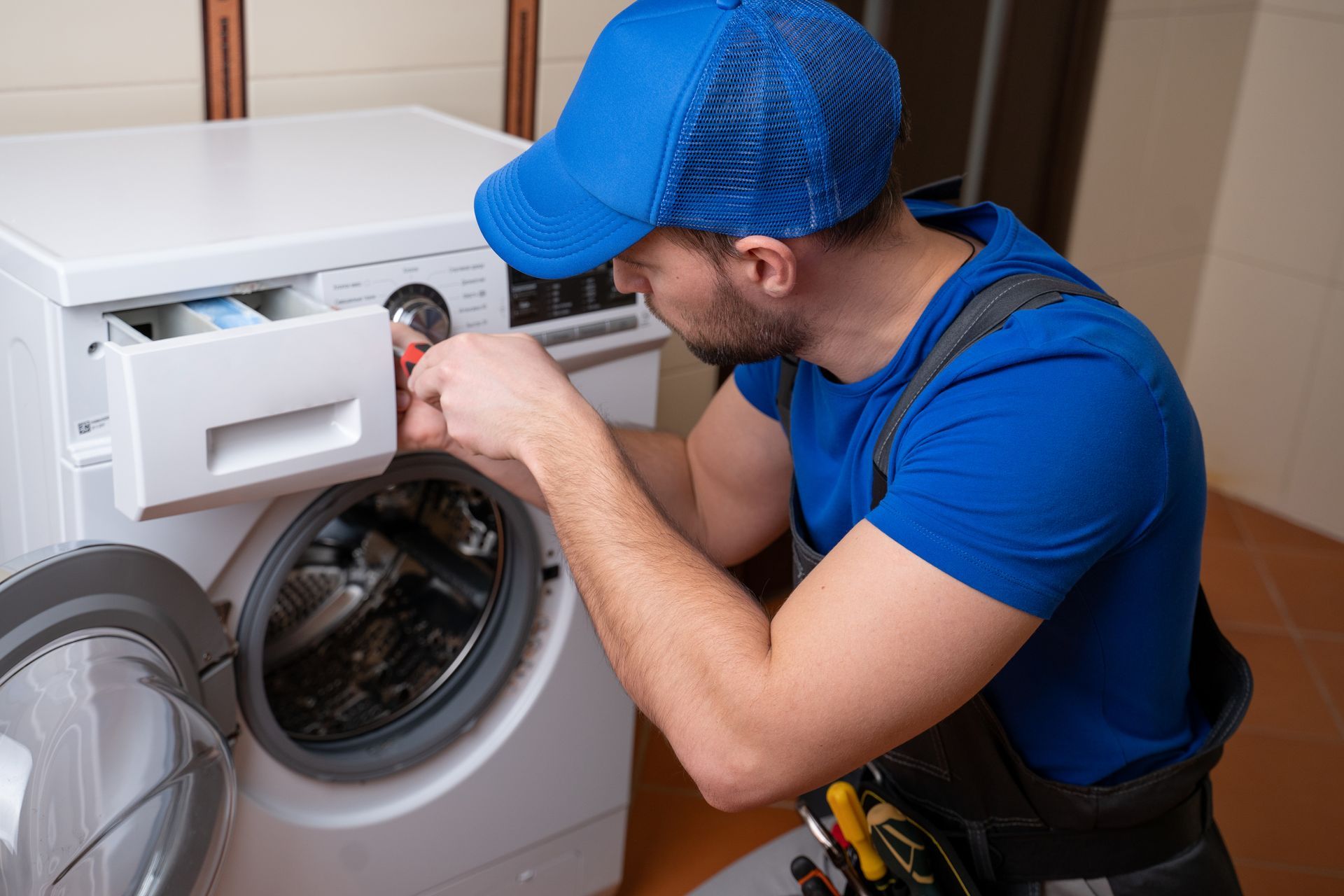 Worker in a blue uniform, performing appliance repair on a washing machine in a laundry room.
