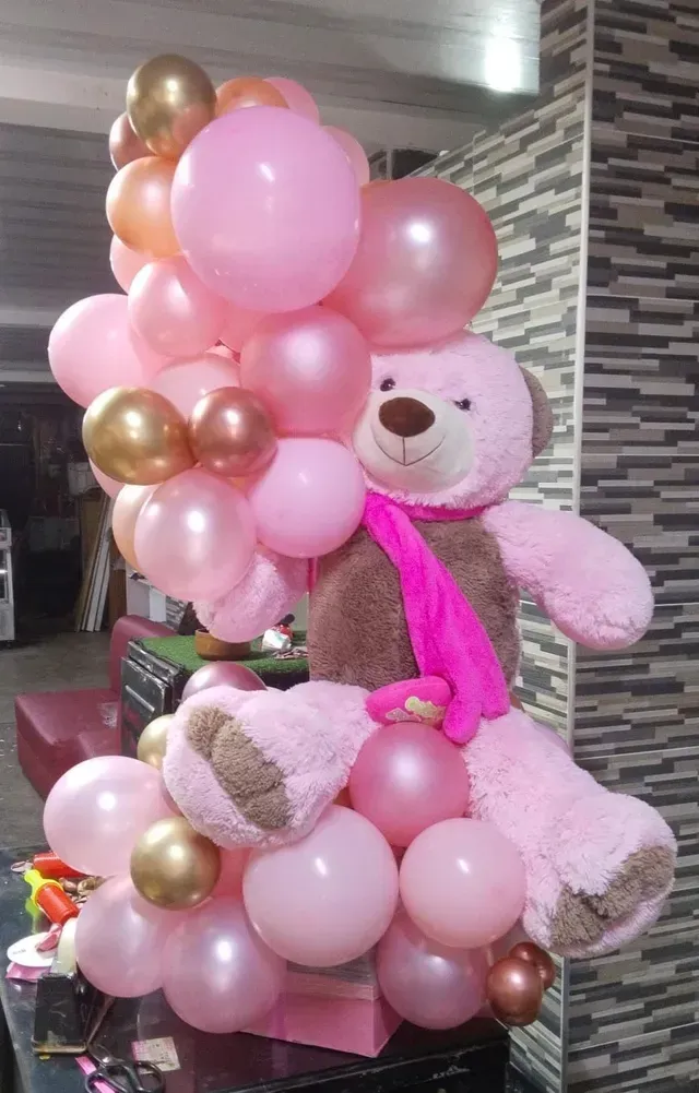 Un osito de peluche rosa rodeado de globos rosas y dorados, sosteniendo una bufanda rosa brillante en el interior.