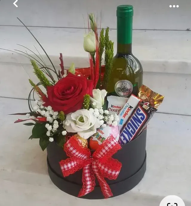 Cesta de regalo con rosas rojas y blancas, botella de vino, barritas de chocolate y cinta en una caja redonda negra.