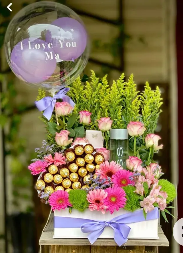 Cesta de regalo floral con globos morados, flores rosas y bombones dorados sobre una mesa blanca.