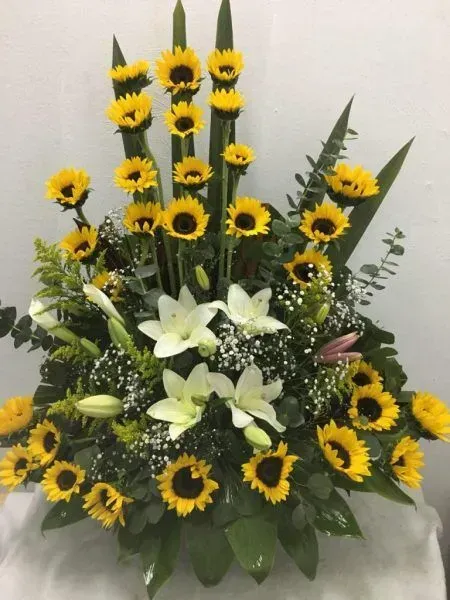 Gran arreglo floral con girasoles, lirios blancos y follaje sobre un mantel blanco.