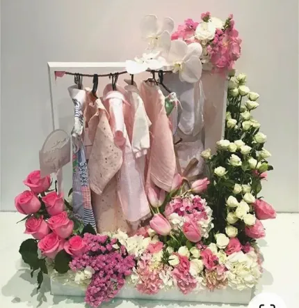 Ropa de bebé en tonos pastel sobre un marco blanco rodeado de flores rosas, blancas y moradas.