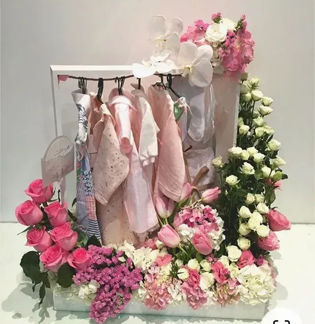 Ropa de bebé en tonos pastel sobre un marco blanco rodeado de flores rosas, blancas y moradas.