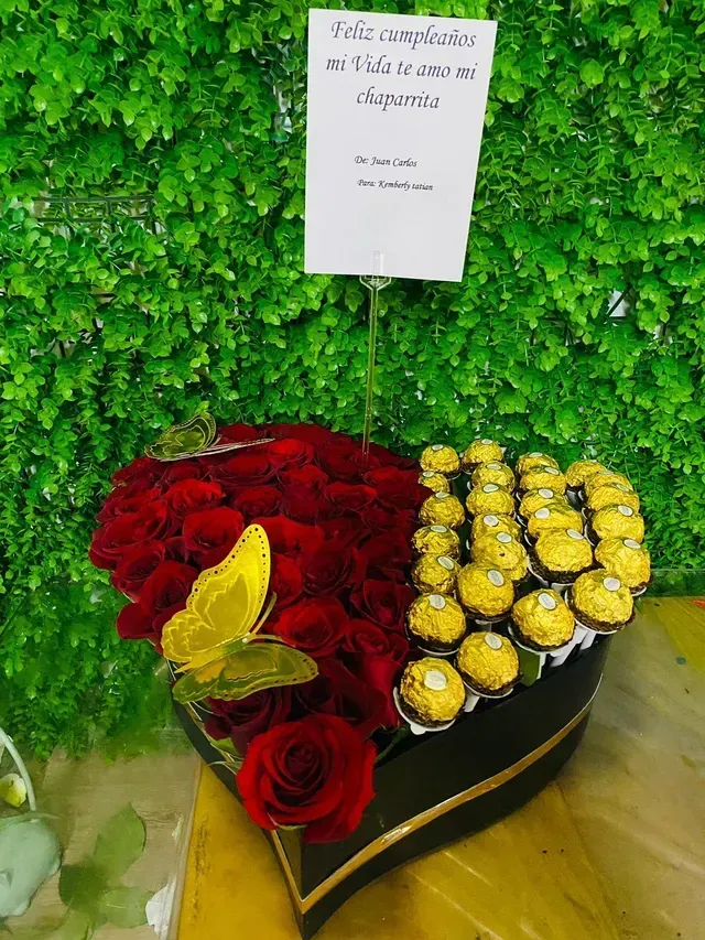 Arreglo de rosas rojas y amarillas en una caja con forma de corazón sobre una pared verde frondosa con una pequeña tarjeta.