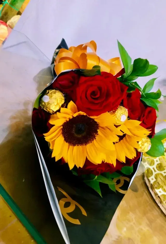 Ramo de rosas rojas, girasoles y flores naranjas envuelto en papel negro sobre una mesa.