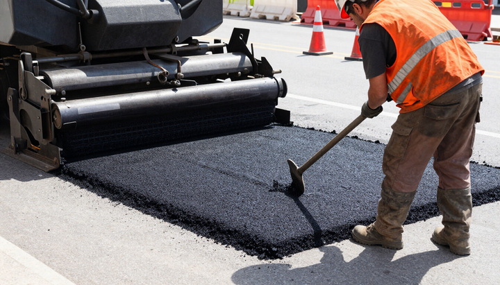 Worker rakes fresh asphalt near a heavy roller during Asphalt Paving Services in Bryan, TX to resurface a major roadway.
