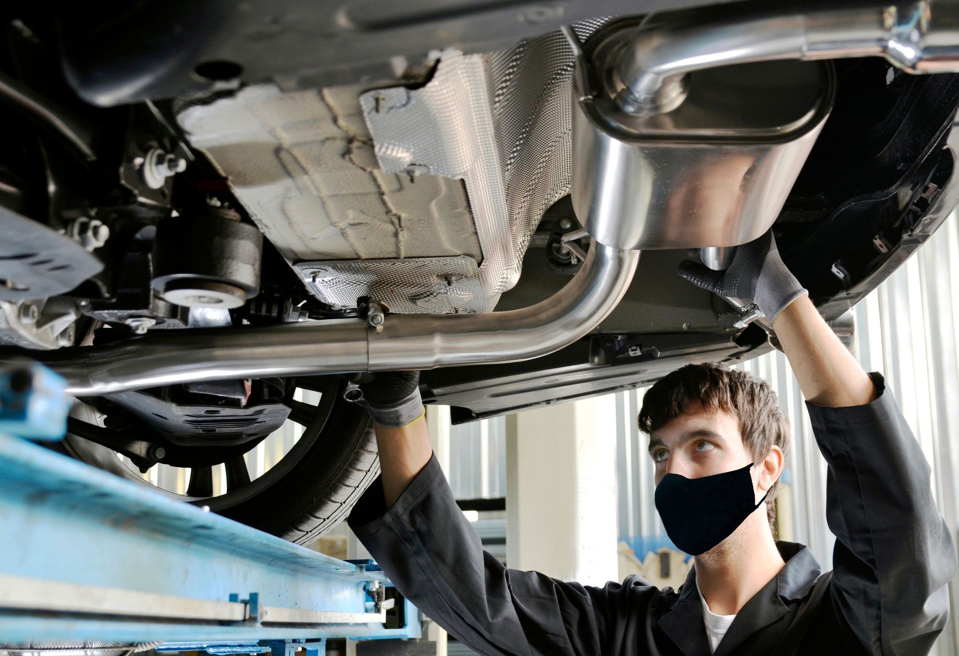 Masked mechanic inspects car’s exhaust and muffler system on lift. Masked mechanic inspects car’s exhaust and muffler system on lift.