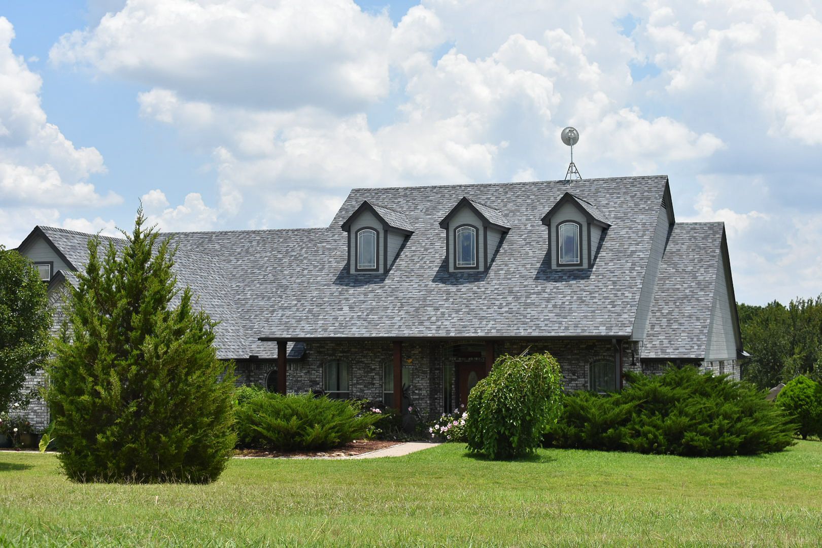 A stone house with a gray shingled roof, three dormer windows, and a front porch, set in a grassy yard under a cloudy sky.