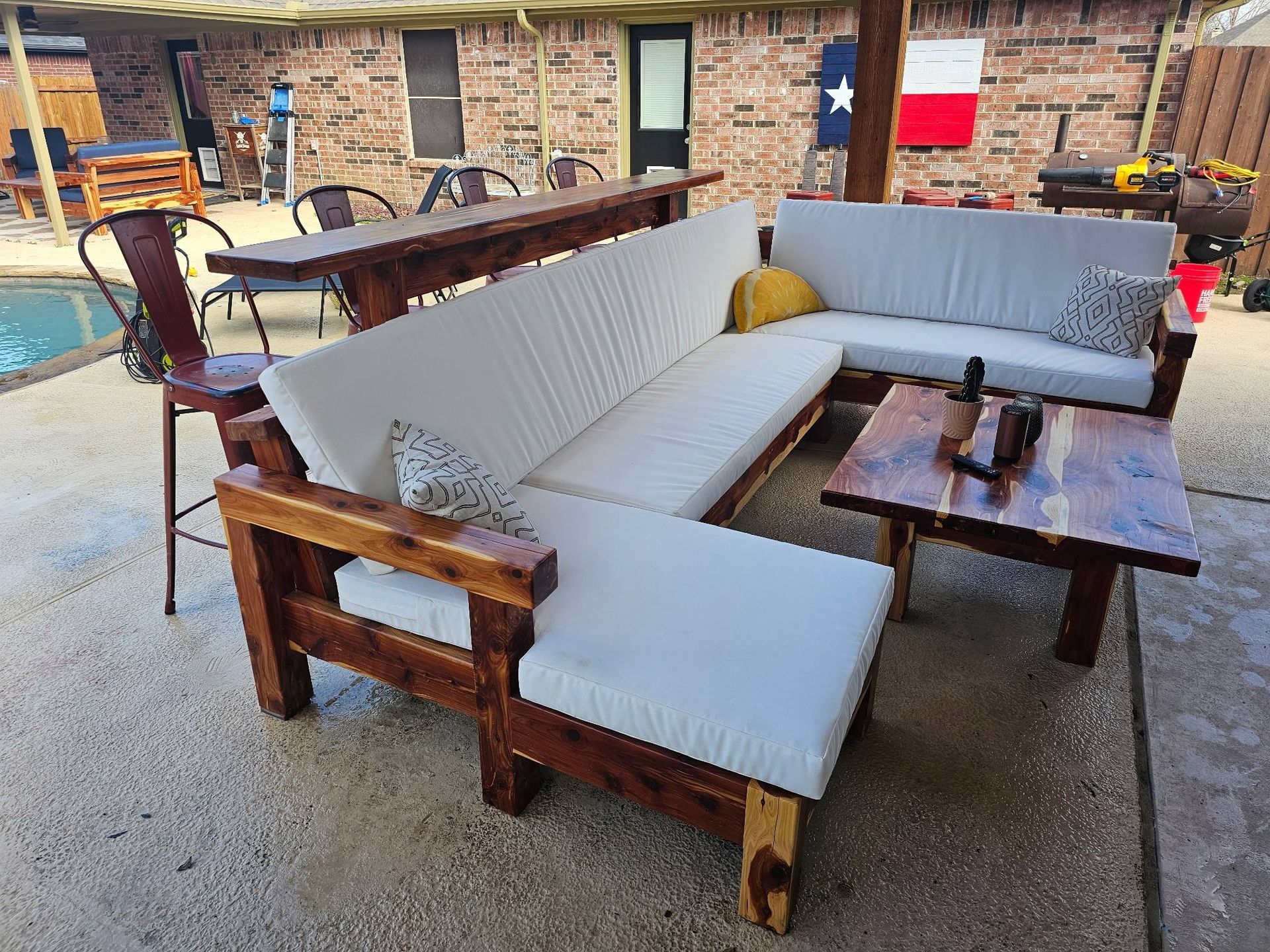 A backyard patio featuring a wooden L-shaped sectional with cream cushions, a matching coffee table, and a bar-height table.