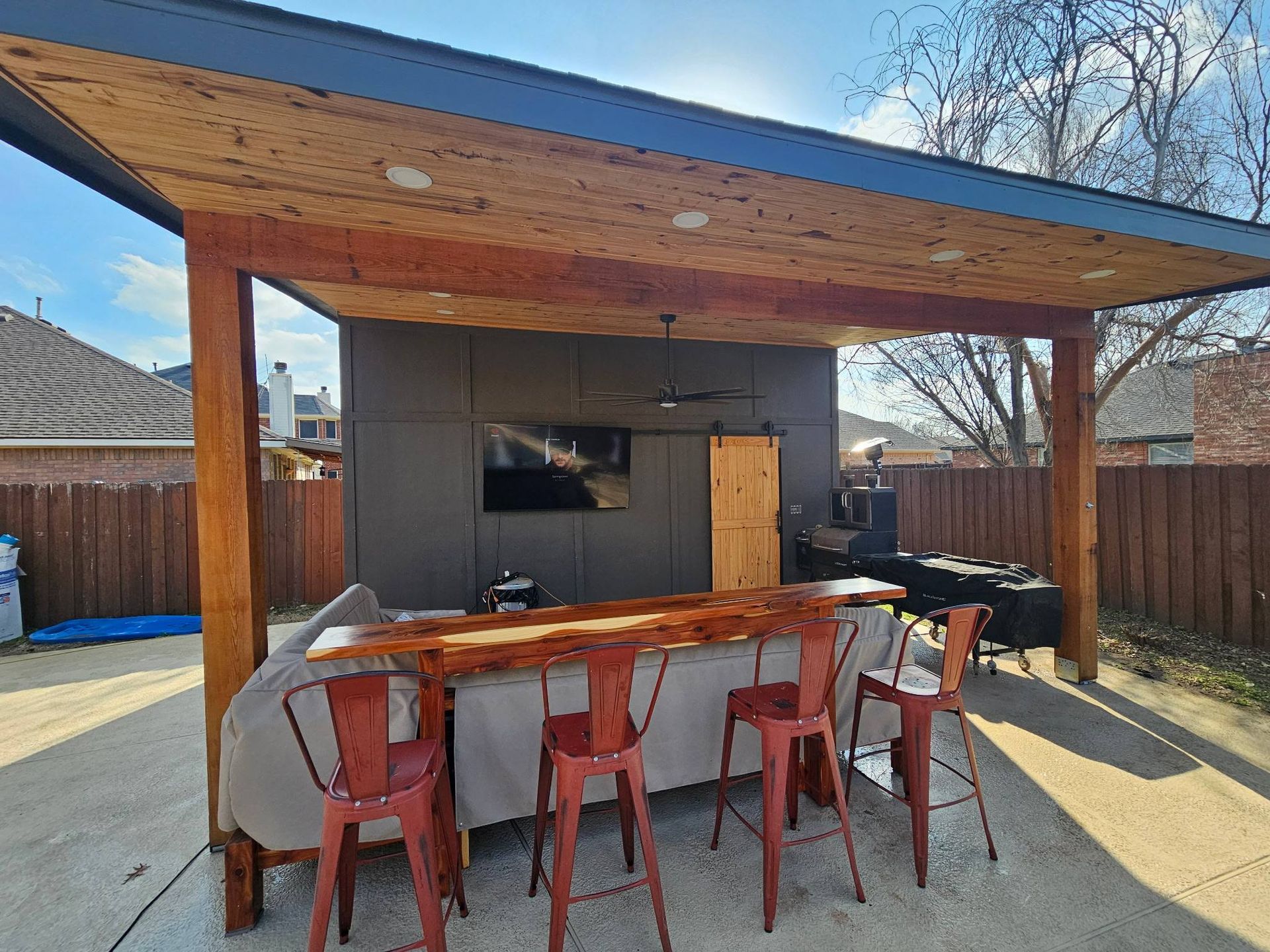A covered outdoor patio with a wooden bar, four red stools, a wall-mounted TV, and a nearby grill.