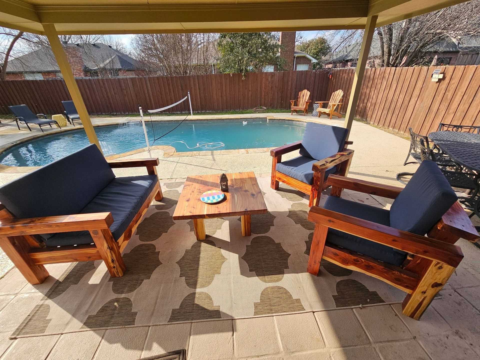 A patio area featuring wooden outdoor furniture with navy blue cushions, a small coffee table, and a pool in the background.