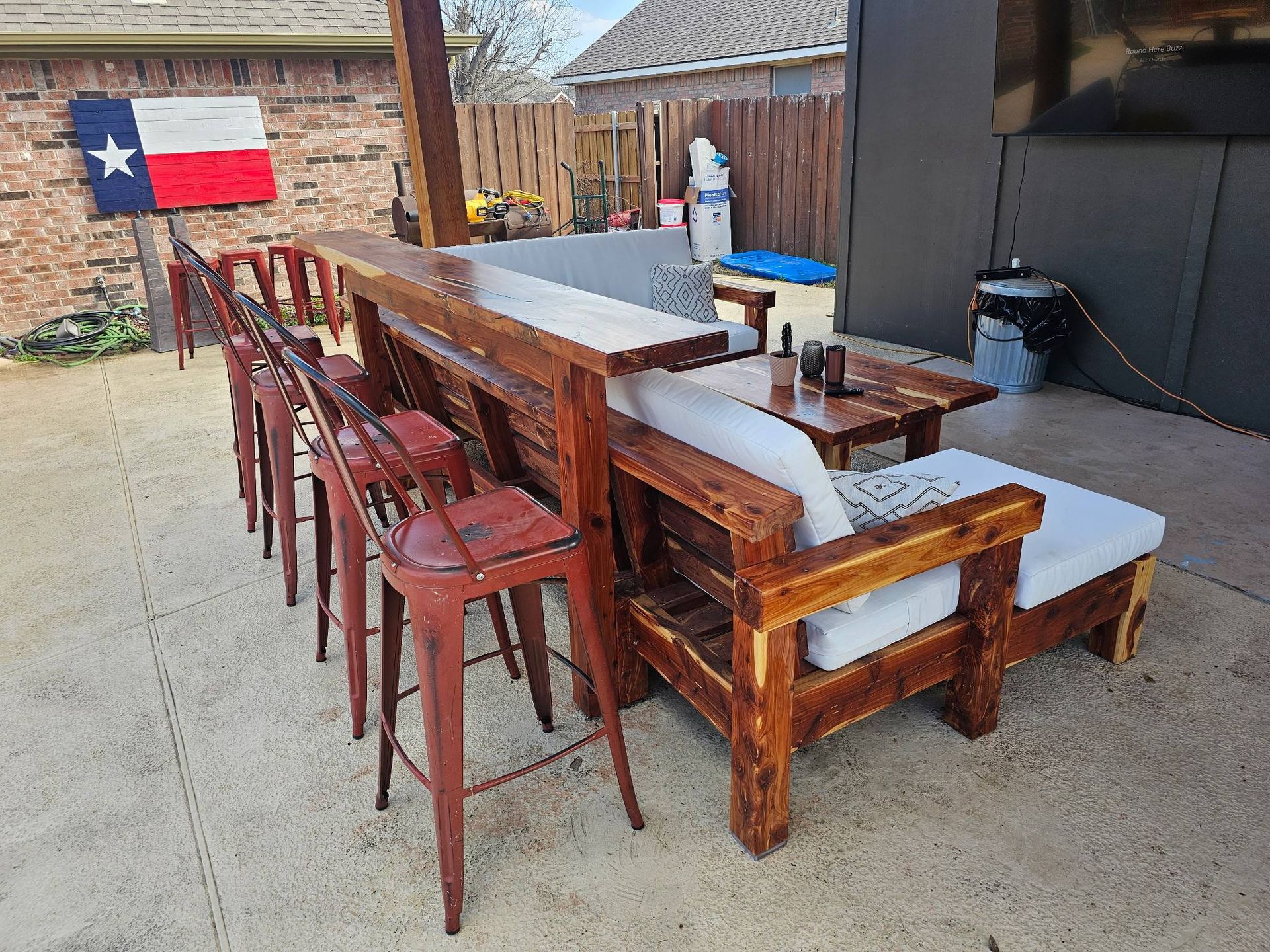 A custom wooden bar set with three red metal stools, cushioned seating, and a Texas flag on a brick patio.