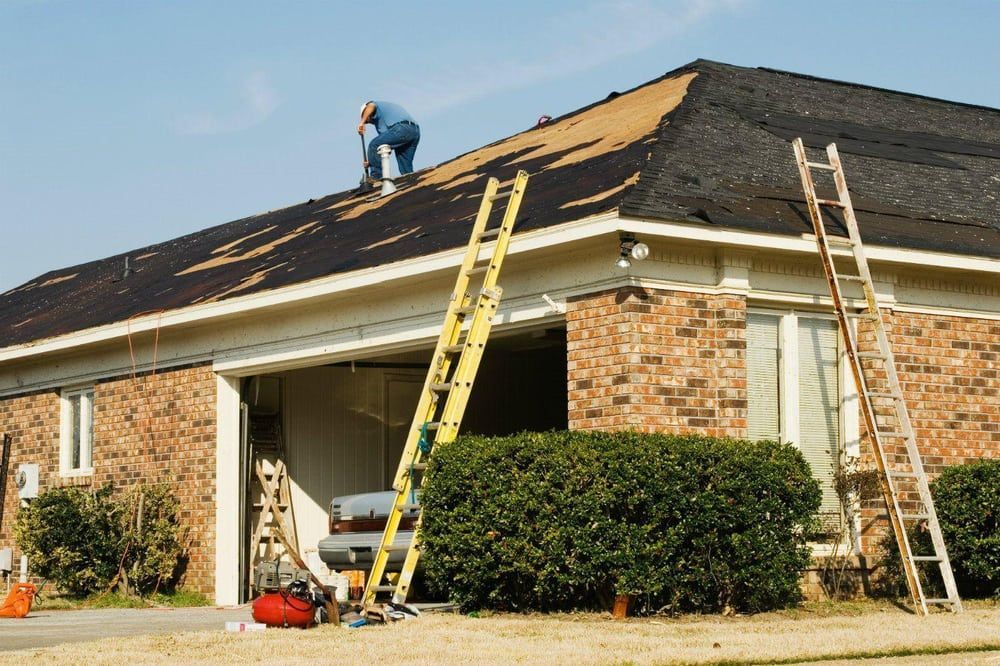 A worker repairing a residential roof with two ladders propped against the brick house and garage.