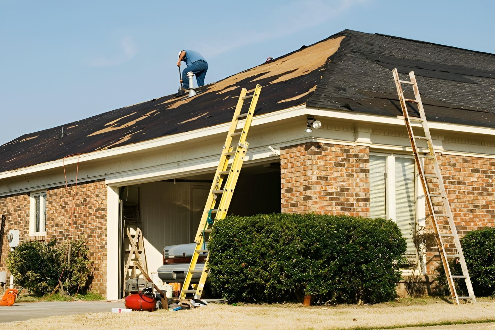 A person working on the dark shingled roof of a brick house, with two tall ladders propped against the building exterior.