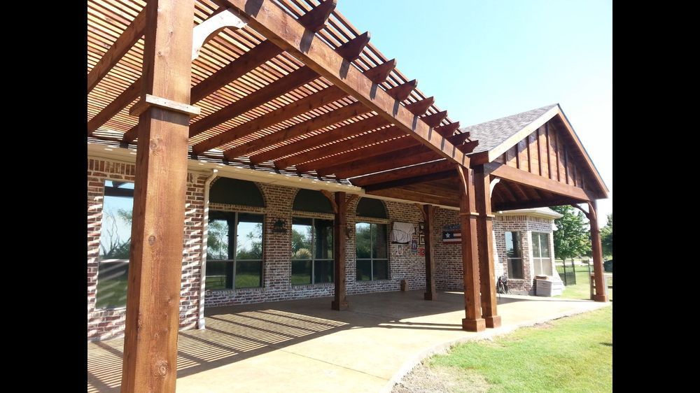 A large wooden pergola extending from a brick house, shading a concrete patio area on a sunny day.