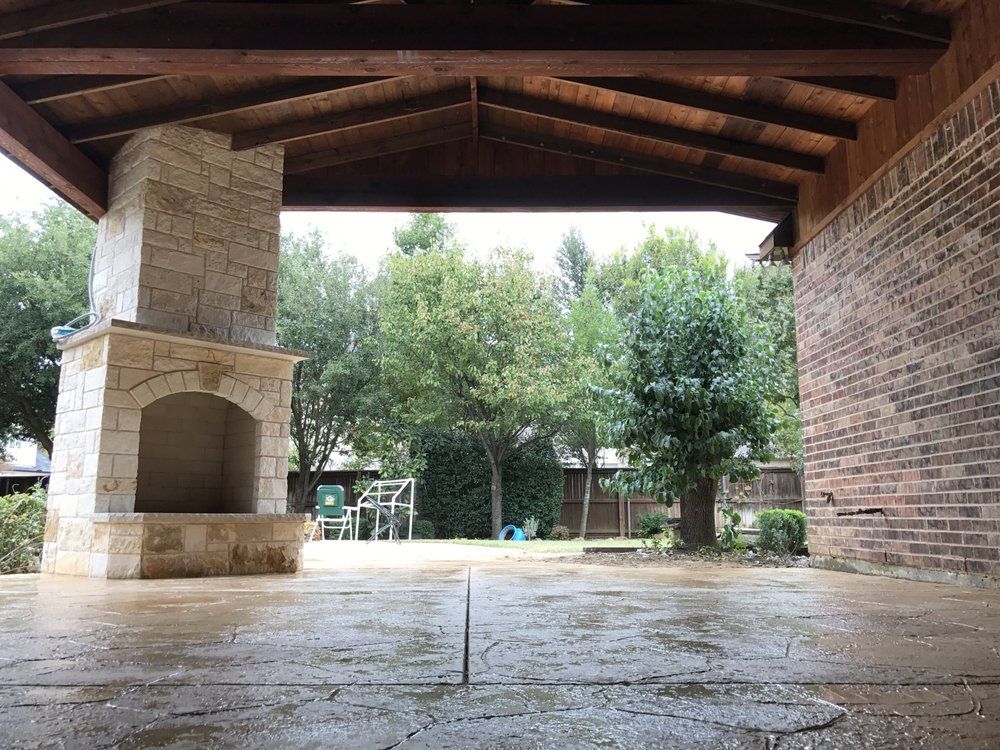 A covered outdoor patio featuring a stone fireplace and a brick wall, looking out onto a backyard with trees.