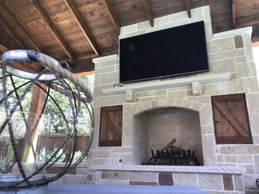 A stone outdoor fireplace with a mounted television above, featuring wooden cabinet doors and a rustic lantern in the foreground.