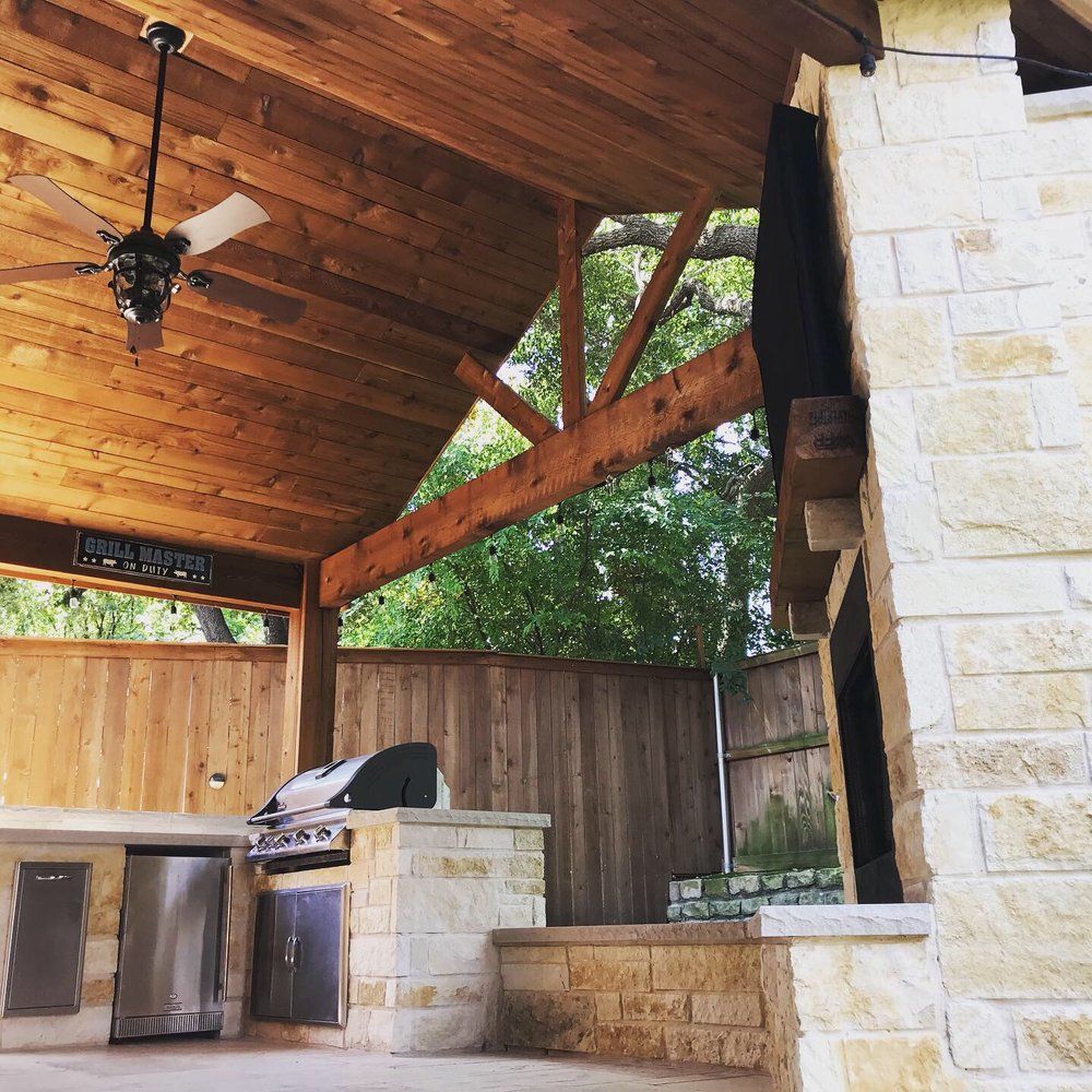 A stone outdoor kitchen with a grill, stainless steel cabinets, and a wood-paneled ceiling under a covered patio.