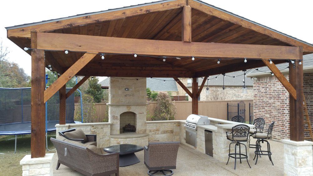 A backyard patio featuring a wood-covered pavilion with an outdoor stone fireplace, built-in grill, and patio furniture.