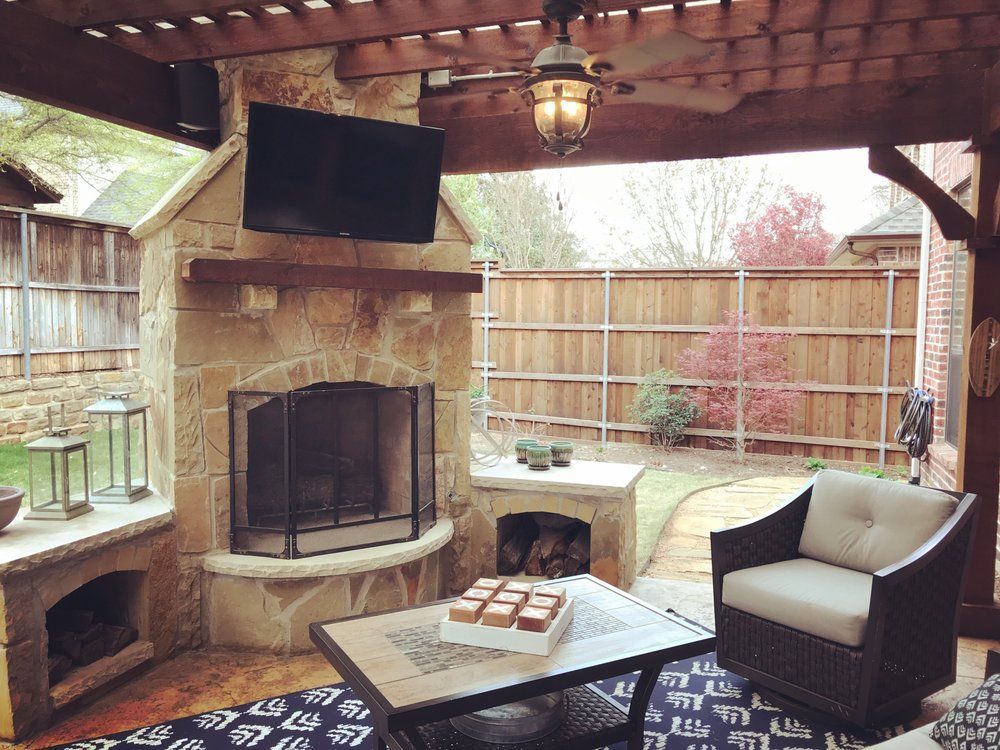 A stone outdoor fireplace with a TV above sits on a patio under a wooden pergola, featuring an armchair and coffee table.