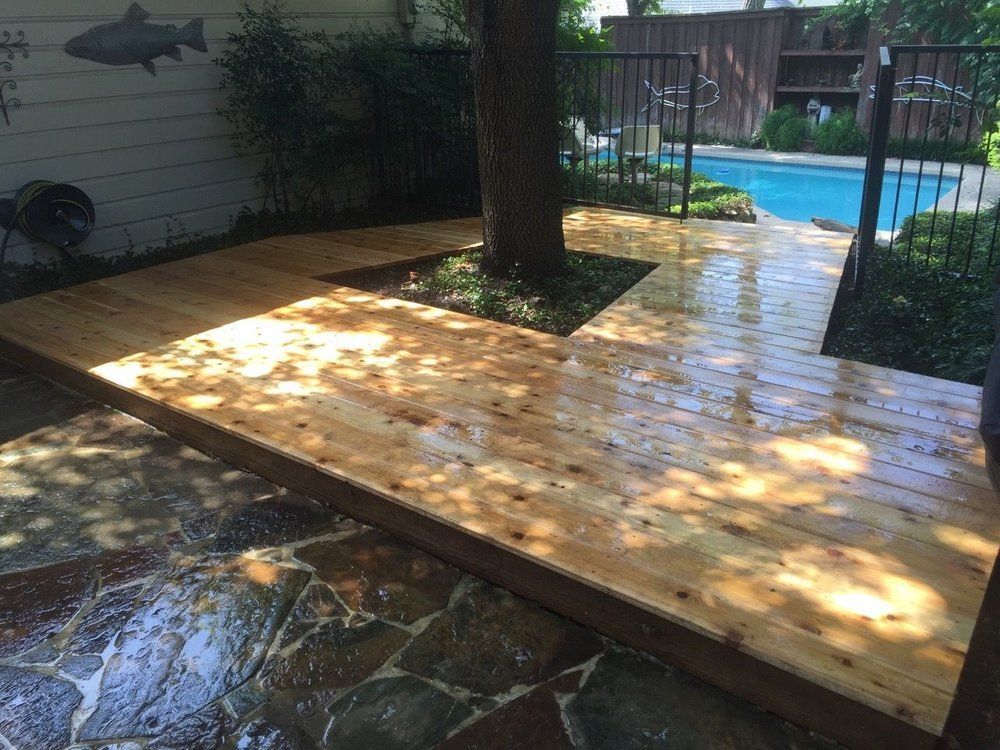 A newly stained wooden deck surrounding a tree, with stone patio flooring in the foreground and a swimming pool beyond.