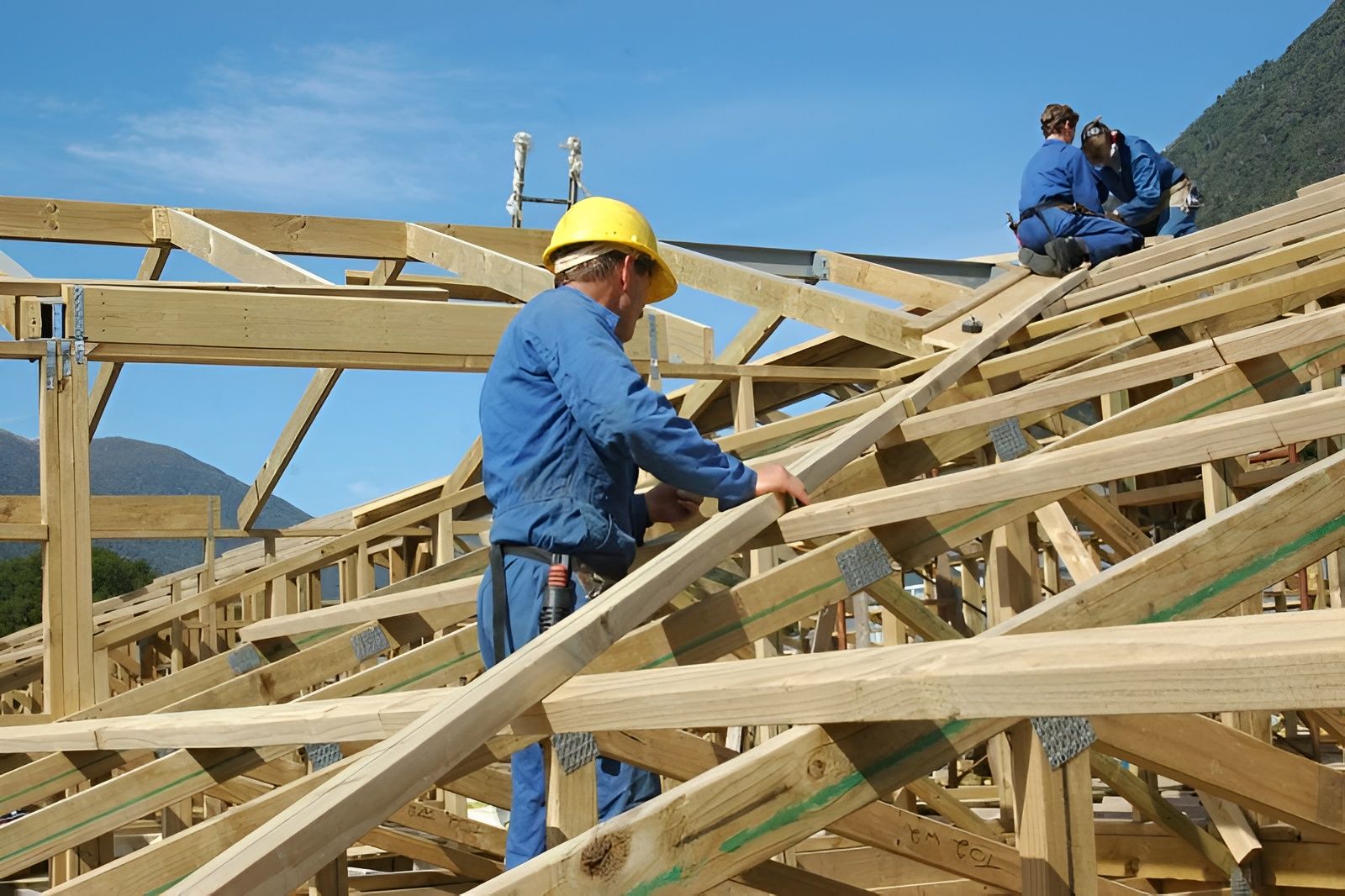 Workers in blue uniforms assemble a wooden roof framework on a building construction site against a clear blue sky.