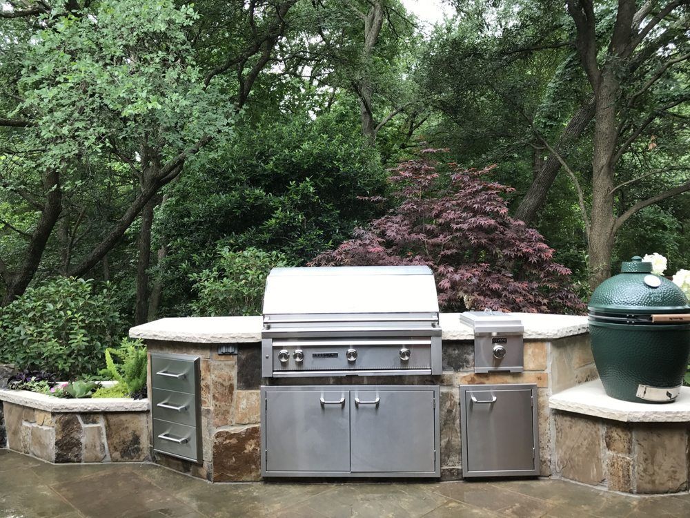 An outdoor kitchen with a stainless steel grill, storage cabinets, and a green ceramic egg-style grill set in stone.