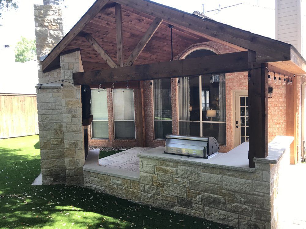 An outdoor patio with a stone fireplace, a covered seating area with wooden beams, and a built-in stone grill island.
