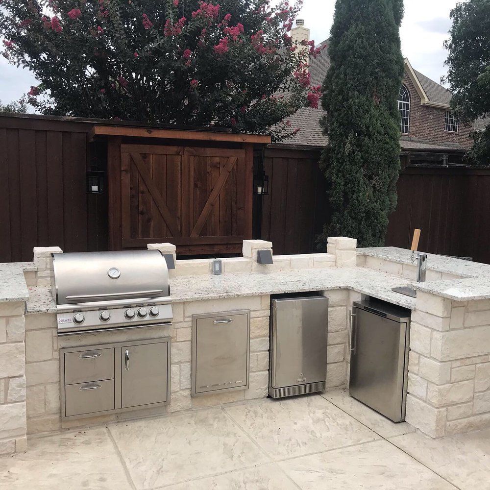 A backyard outdoor kitchen with a stone-clad L-shaped counter, built-in stainless grill, cabinetry, and a small sink.