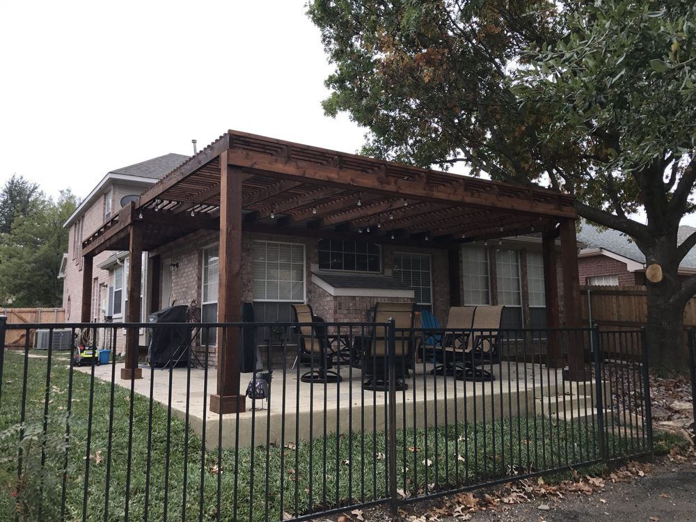 A dark wood pergola covers a concrete patio with outdoor furniture, viewed through a black metal fence in a backyard.