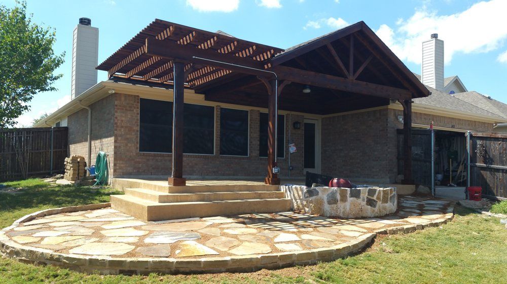 A backyard patio featuring a stone flagstone floor, concrete steps, and a dark wooden pergola attached to a brick house.
