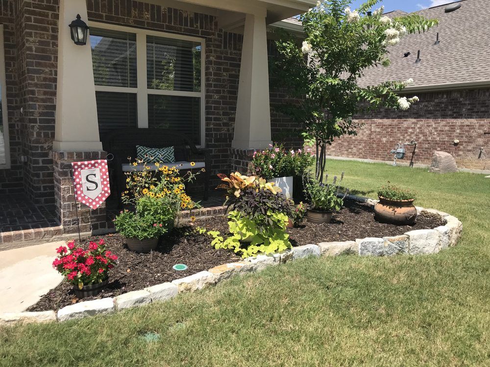 A brick home front with a landscaped garden bed containing colorful flowers, shrubs, and a decorative stone border.
