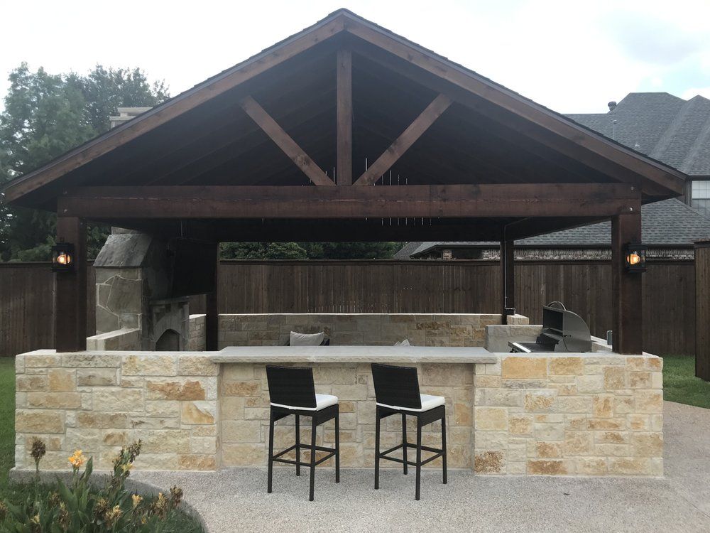 An outdoor kitchen with a stone bar, two stools, a grill, and a stone oven under a wooden pavilion in a fenced yard.
