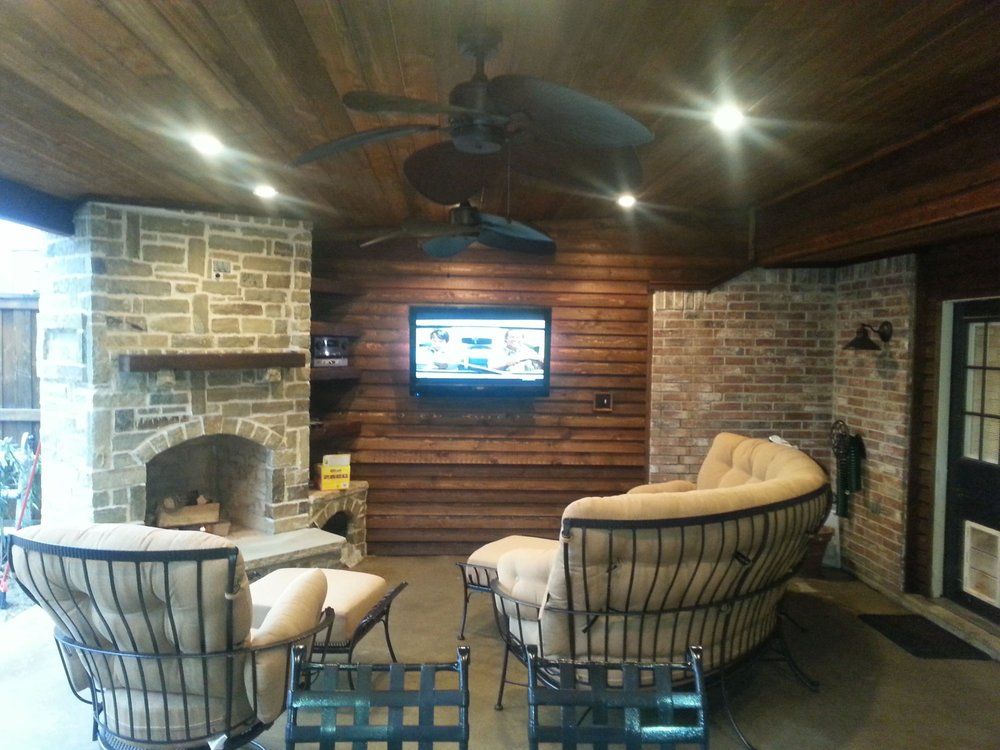 A covered outdoor patio with a stone fireplace, wood-paneled walls, a wall-mounted TV, and lounge furniture.