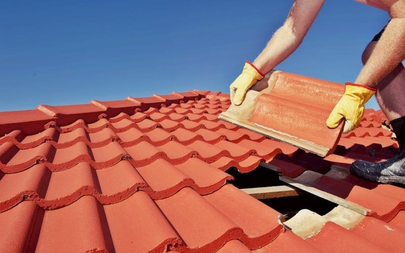 A roofer in yellow gloves installs a red clay roof tile onto a house roof against a clear blue sky.