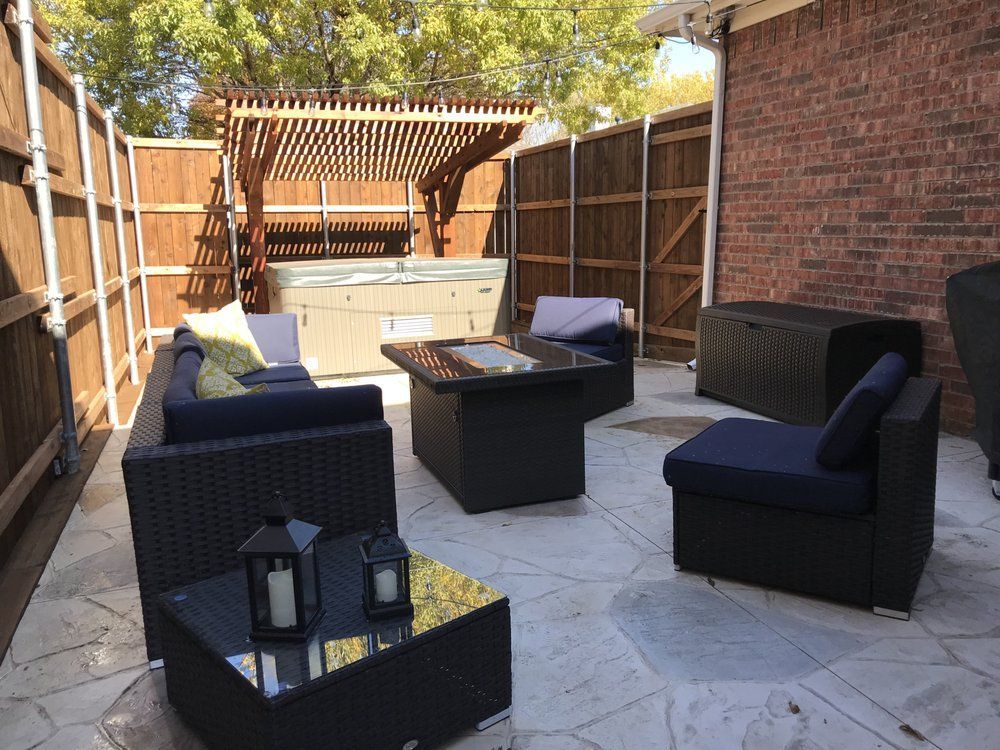 A patio with dark wicker outdoor furniture, a fire pit table, and a hot tub under a wooden pergola, enclosed by a fence.
