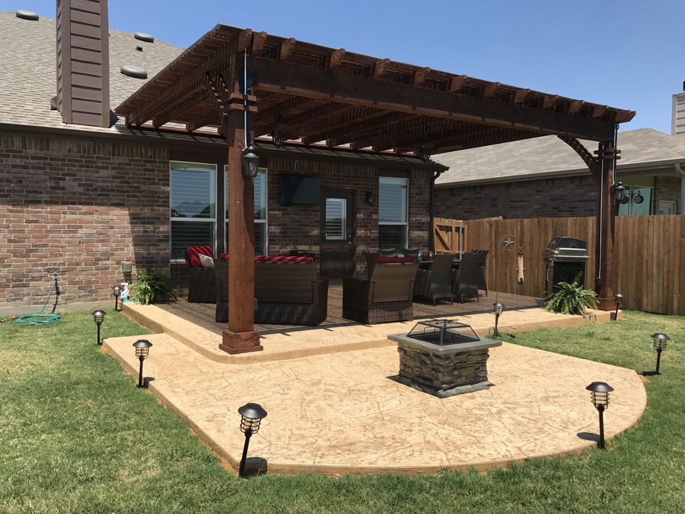 A tan concrete patio with a wooden pergola, outdoor seating, and a stone fire pit in a residential backyard.