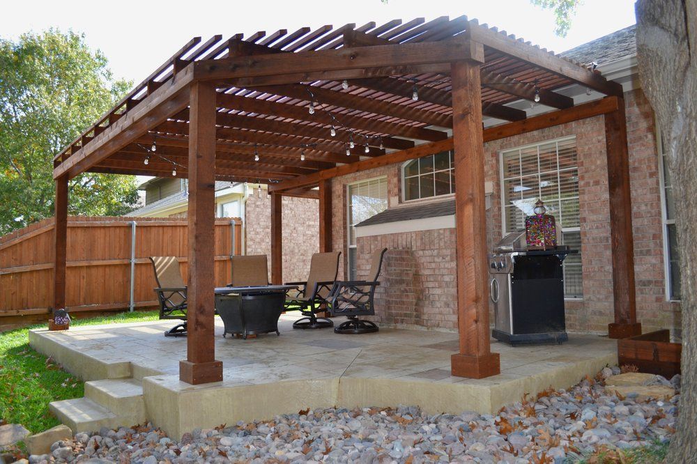 A wooden pergola with string lights over a stone patio featuring patio chairs, a fire pit, and a grill beside a brick house.