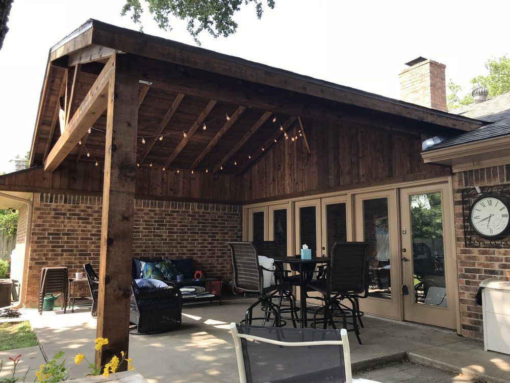 Covered patio with wooden rafters and string lights, featuring outdoor furniture against a brick house wall.