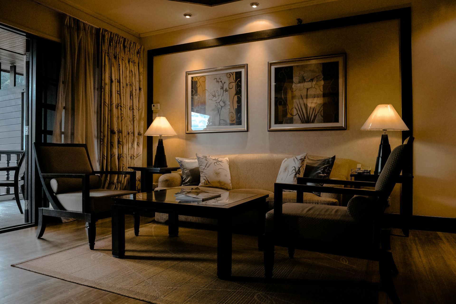 A cozy, warmly lit hotel sitting area with a sofa, two wooden chairs, a coffee table, and two lamps beneath framed art.
