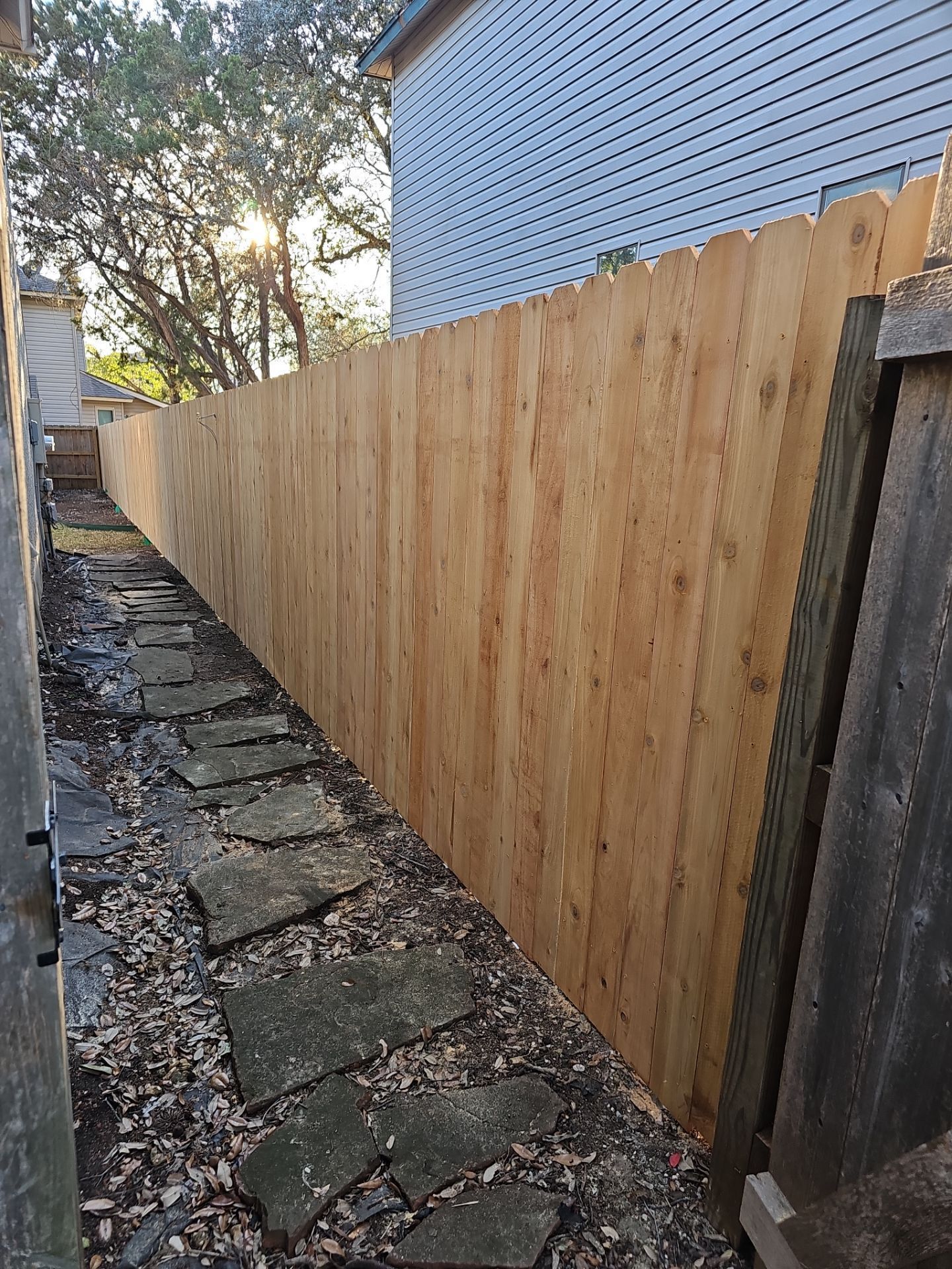 A narrow outdoor pathway with stone steps runs alongside a tall, new wooden fence. The fence extends into the distance.