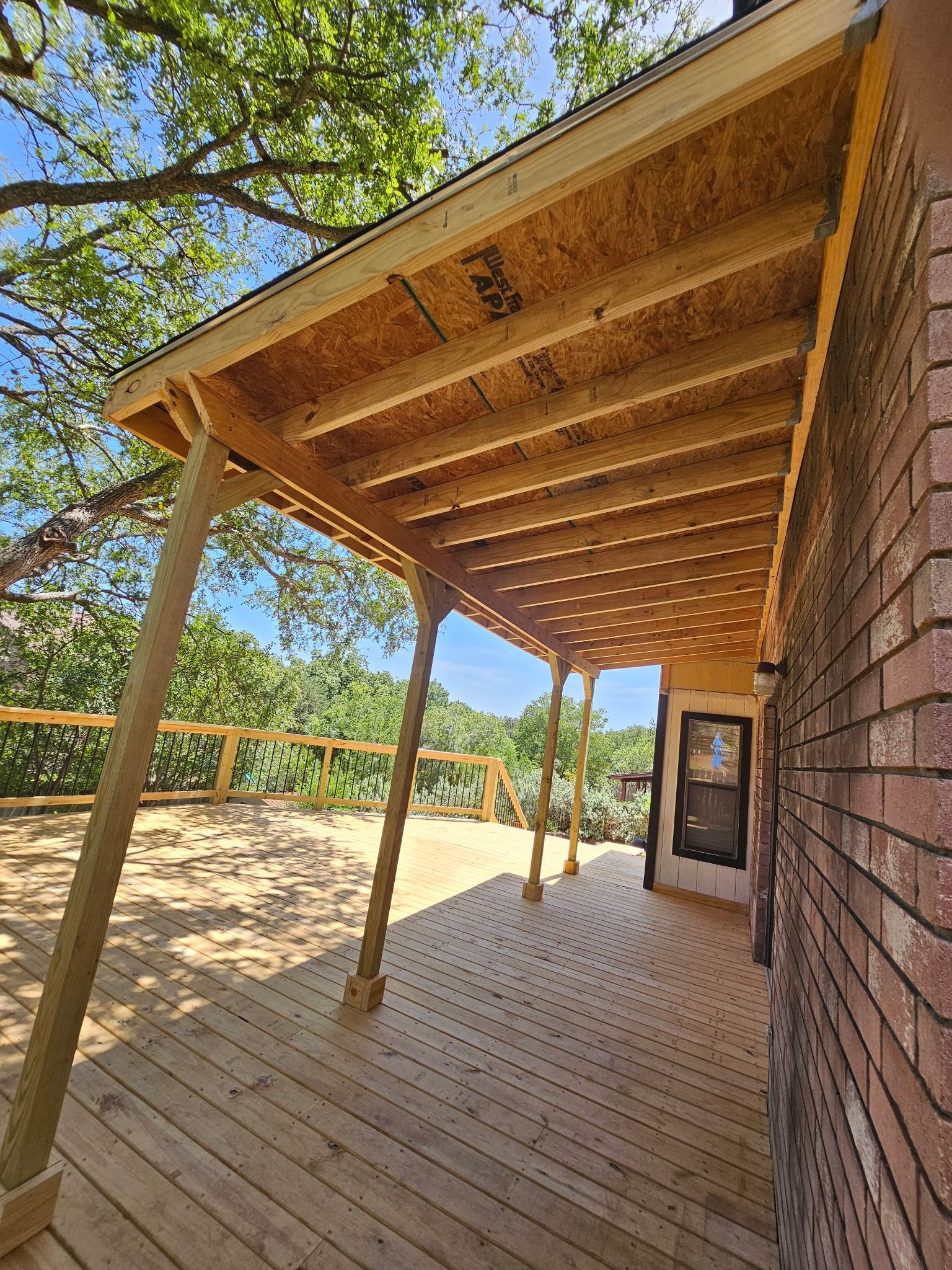 Wooden deck with attached roof supported by posts. Brick house on right, trees and sky visible.