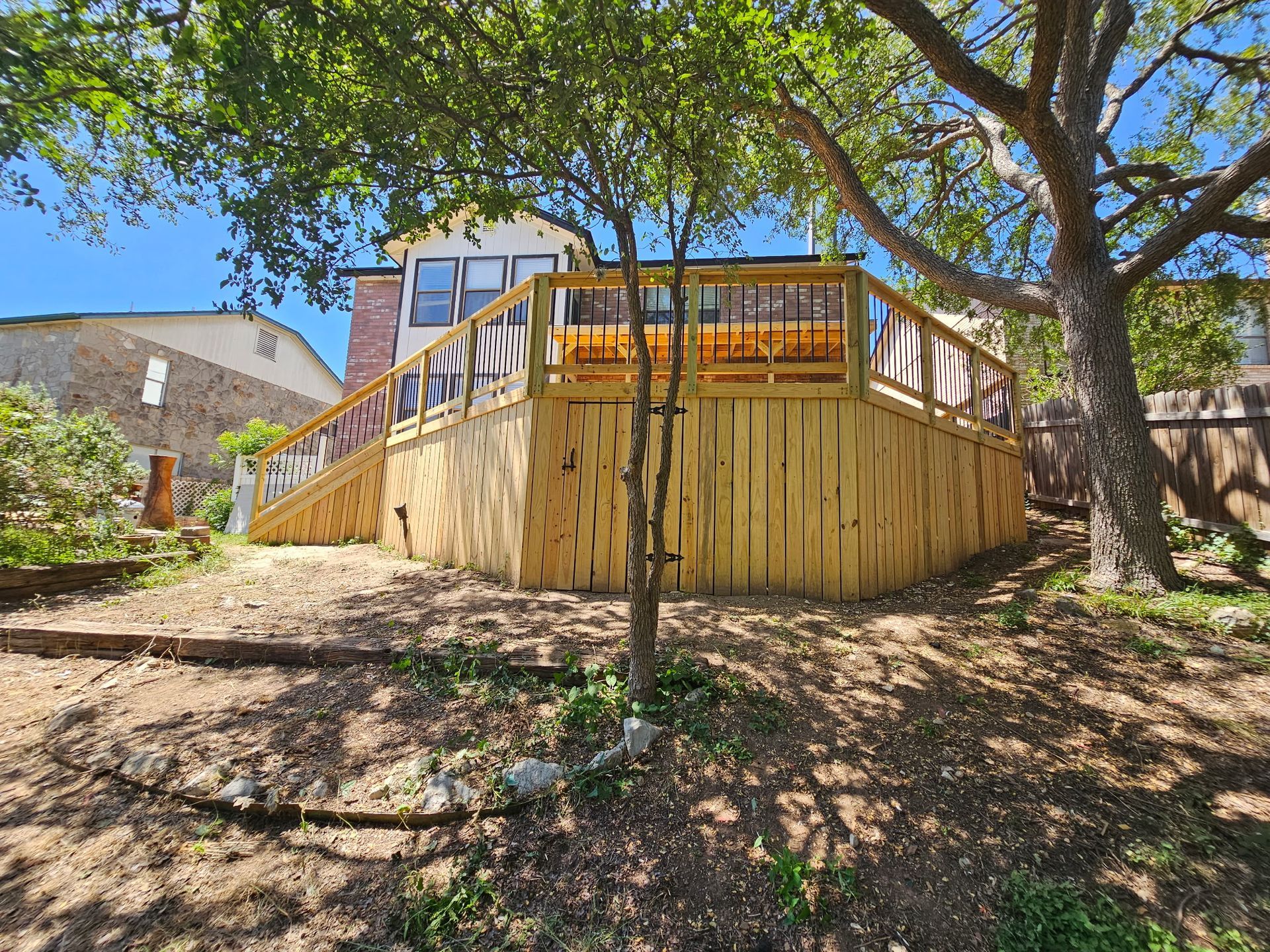 Newly built wooden deck with clear railings, next to a two-story house, under a sunny, tree-filled sky.