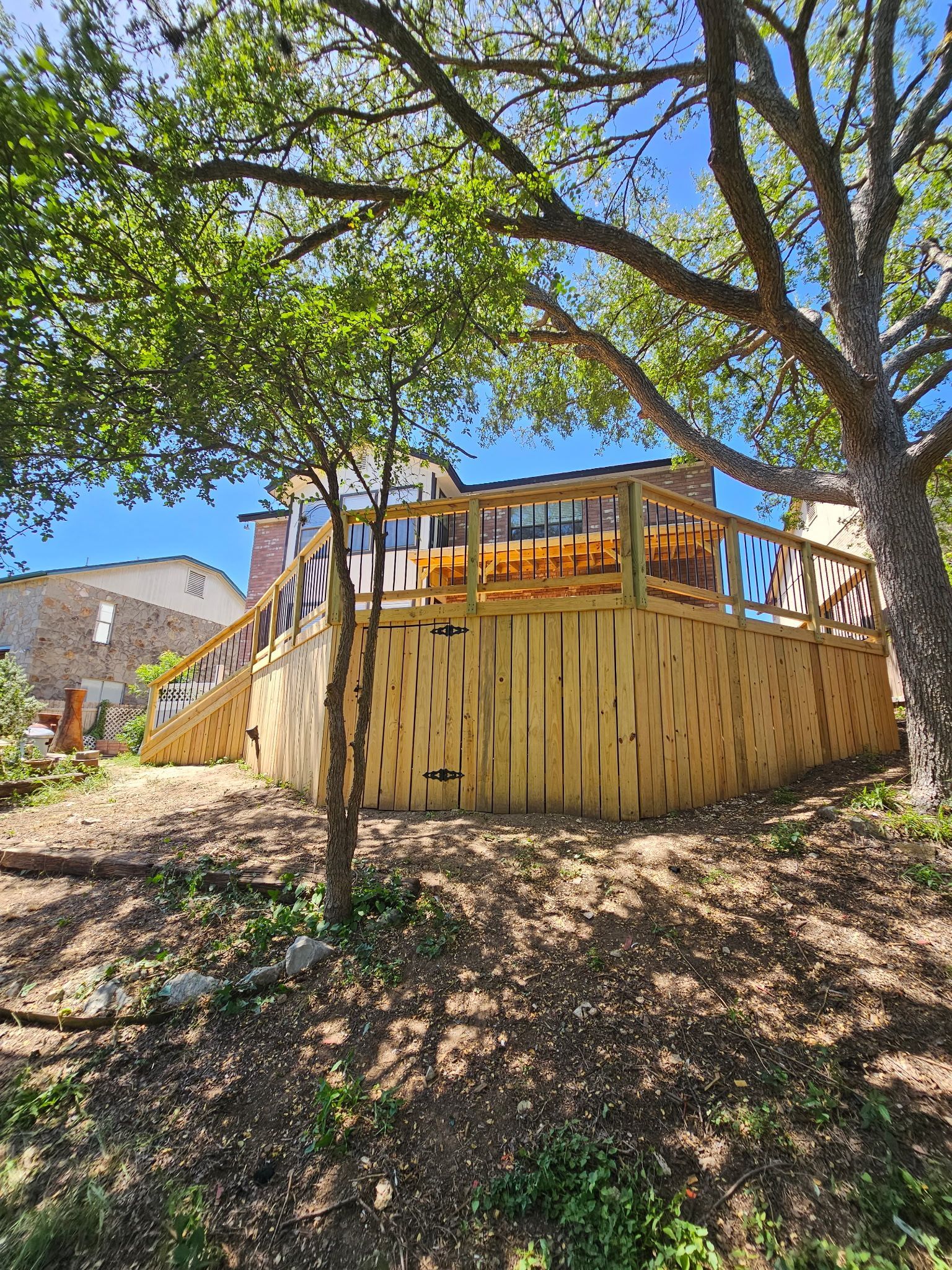 Wooden deck with railings in a backyard setting, surrounded by trees, with a house in the background.
