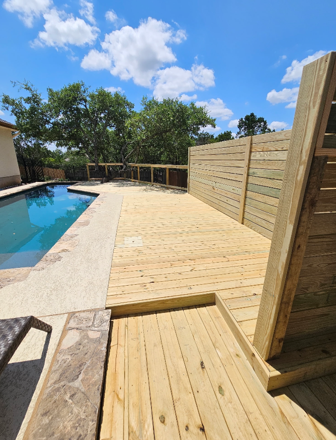 Wooden deck next to a swimming pool, with a privacy fence. Sunny day, blue sky, and green trees in the background.