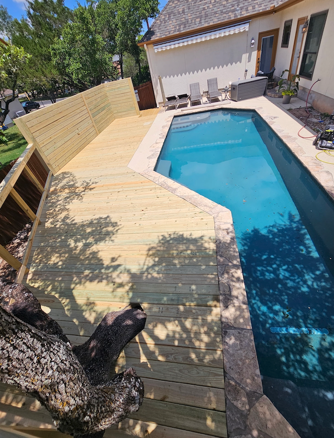 Wooden deck next to a swimming pool. Deck is light brown; pool is turquoise. Sun shade hangs from the side of the house.