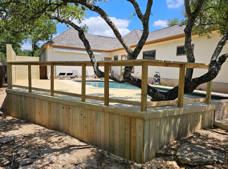 Wooden deck with railing surrounds a swimming pool, built against a beige house with a tree branch overhead.